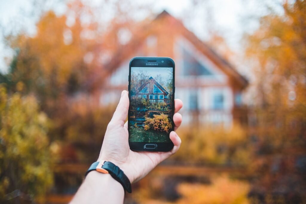 A hand holds a smartphone to photograph a house framed by autumn colors.