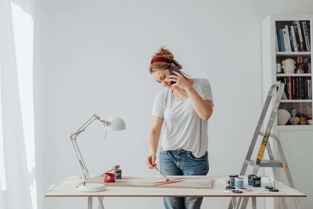 Caucasian woman multitasking with paintbrush and phone at home.