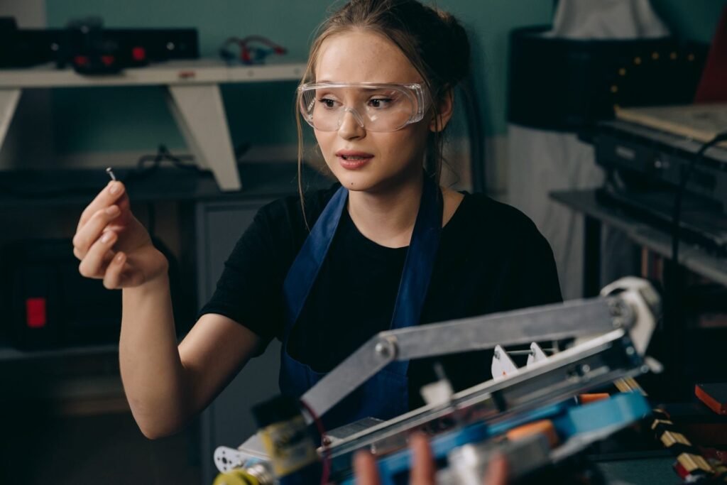 Caucasian woman wearing protective eyewear inspects robotic machinery part indoors.